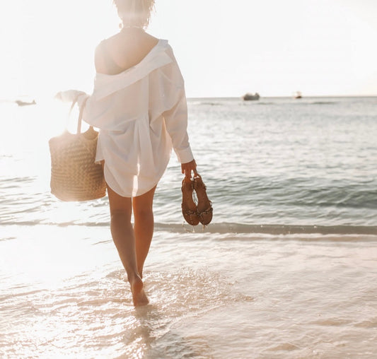 woman standing on beach, facing ocean, holding sandals and tote bag