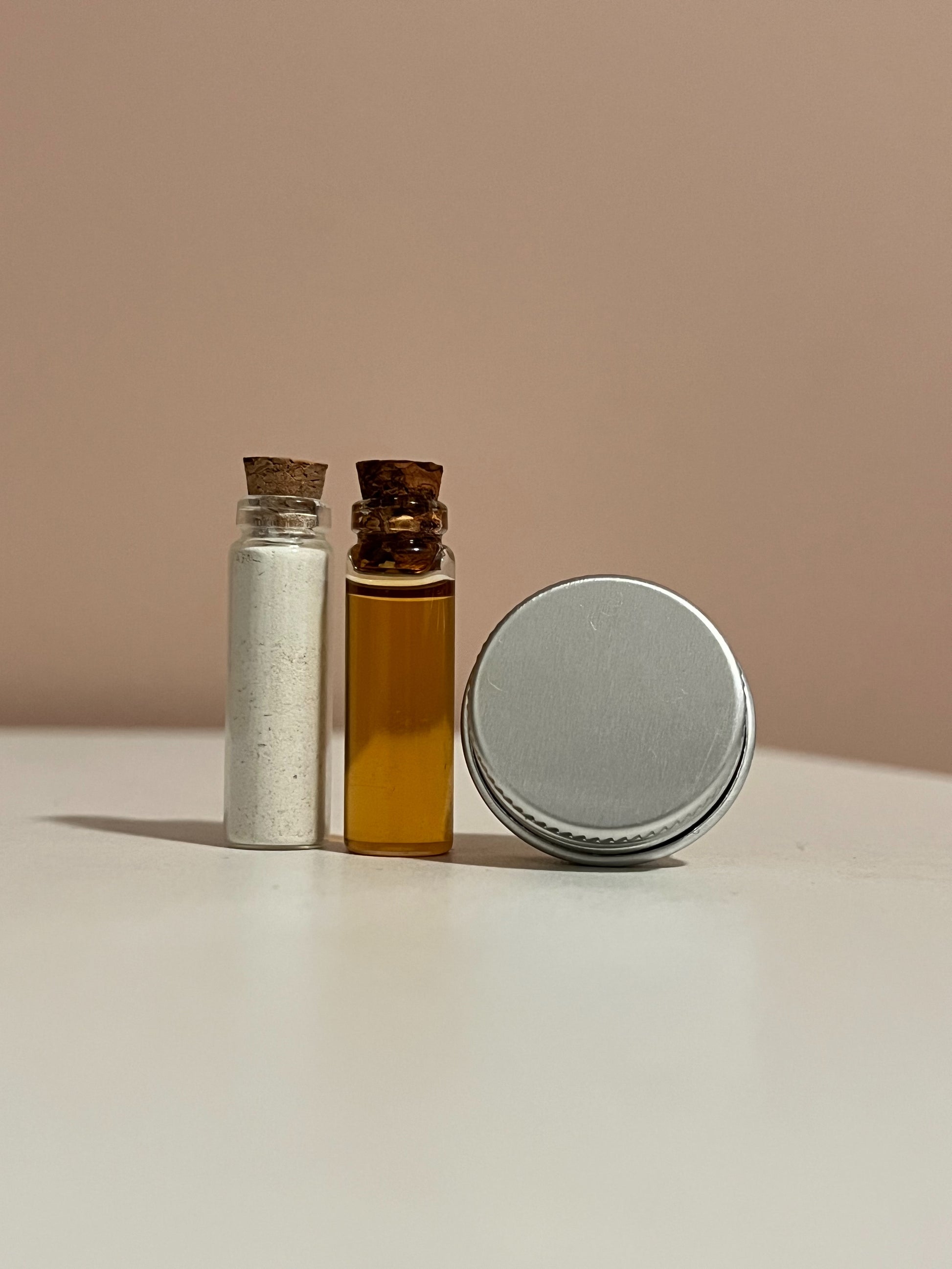 2g glass vial filled with powdered cleanser beside 2g glass vial filled with facial oil serum beside 5g silver tin filled with balm