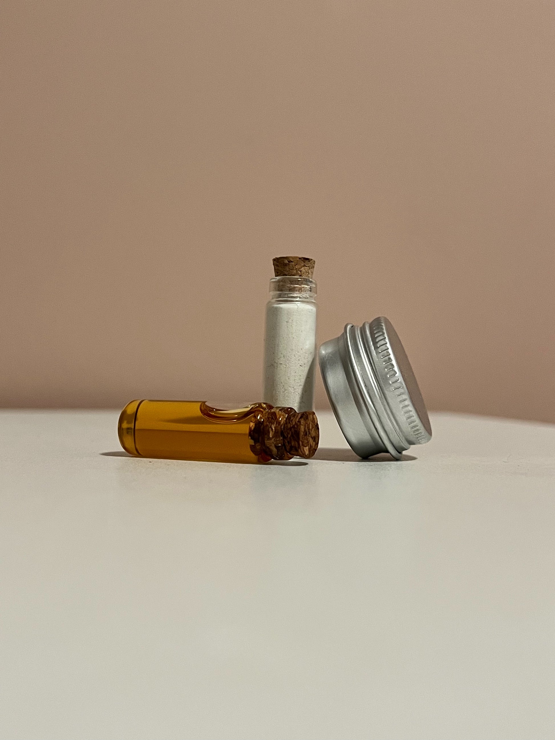 2g glass vial filled with powdered cleanser standing upright with 2g glass vial filled with facial oil serum on it's side and 5g silver tin filled with balm tilted diagonally
