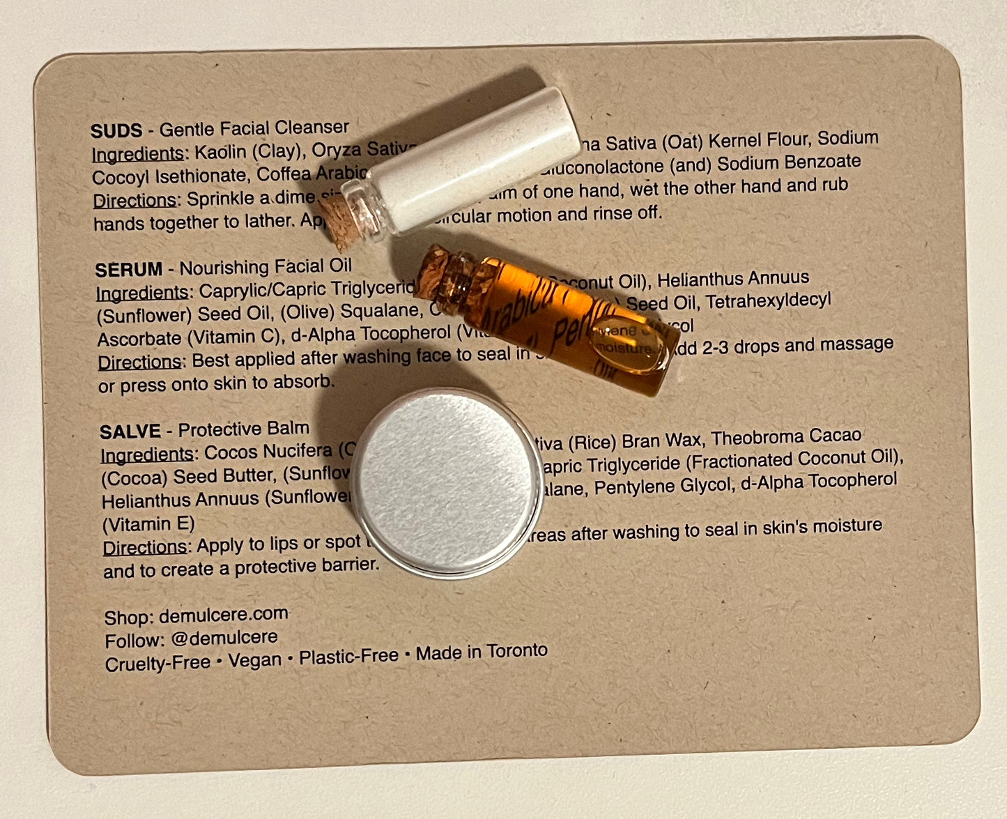 2g glass vial filled with powdered cleanser, 2g glass vial filled with facial oil serum, and 5g silver tin filled with balm all laying flat on top of typed product descriptions with ingredients and directions