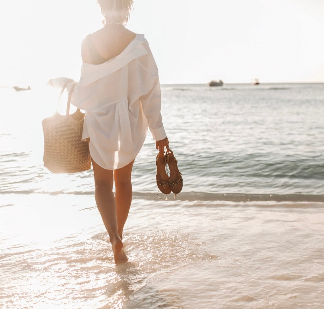 woman standing on beach, facing ocean, holding sandals and tote bag