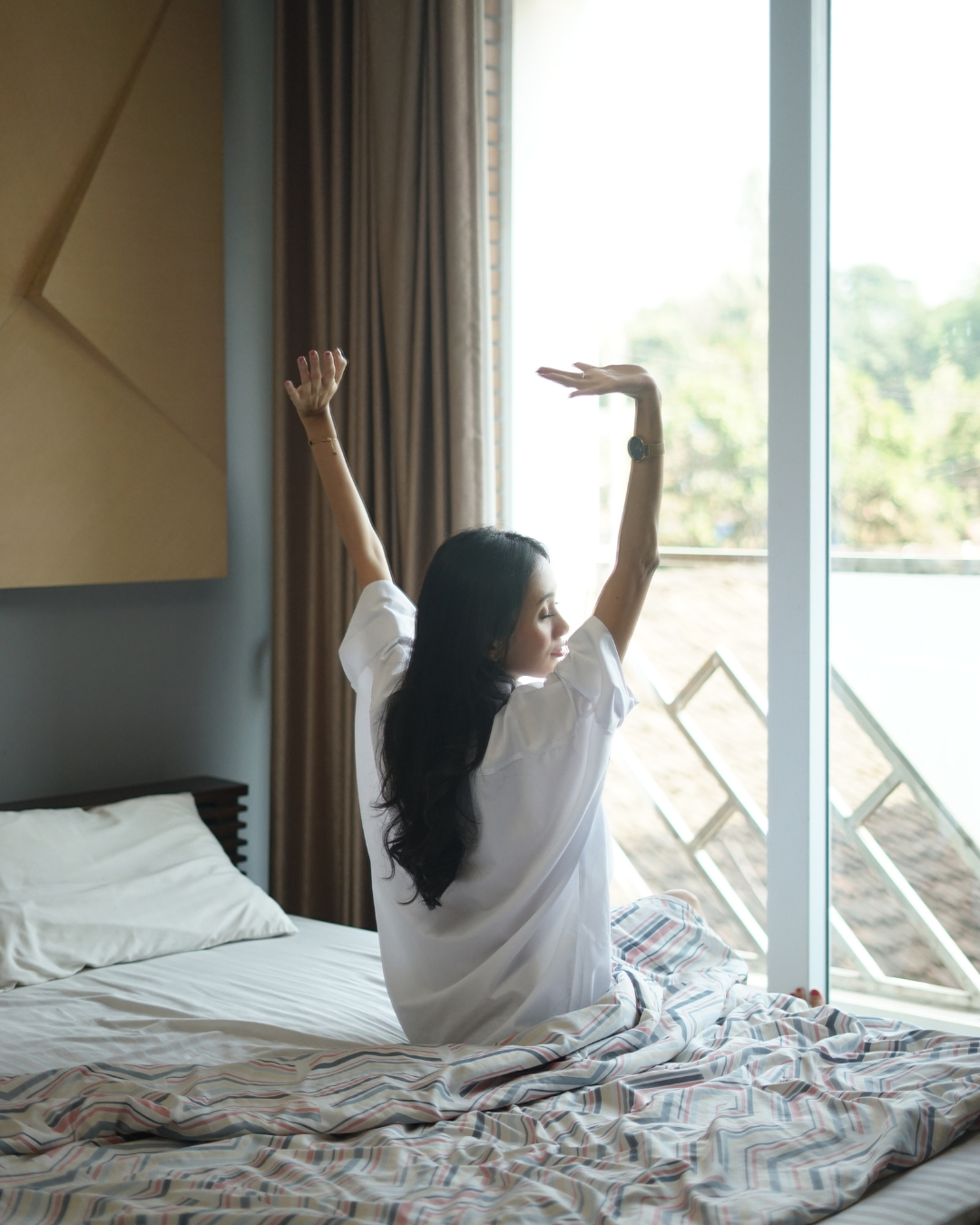 woman sitting on be stretching her arms up in front of a large sunny window