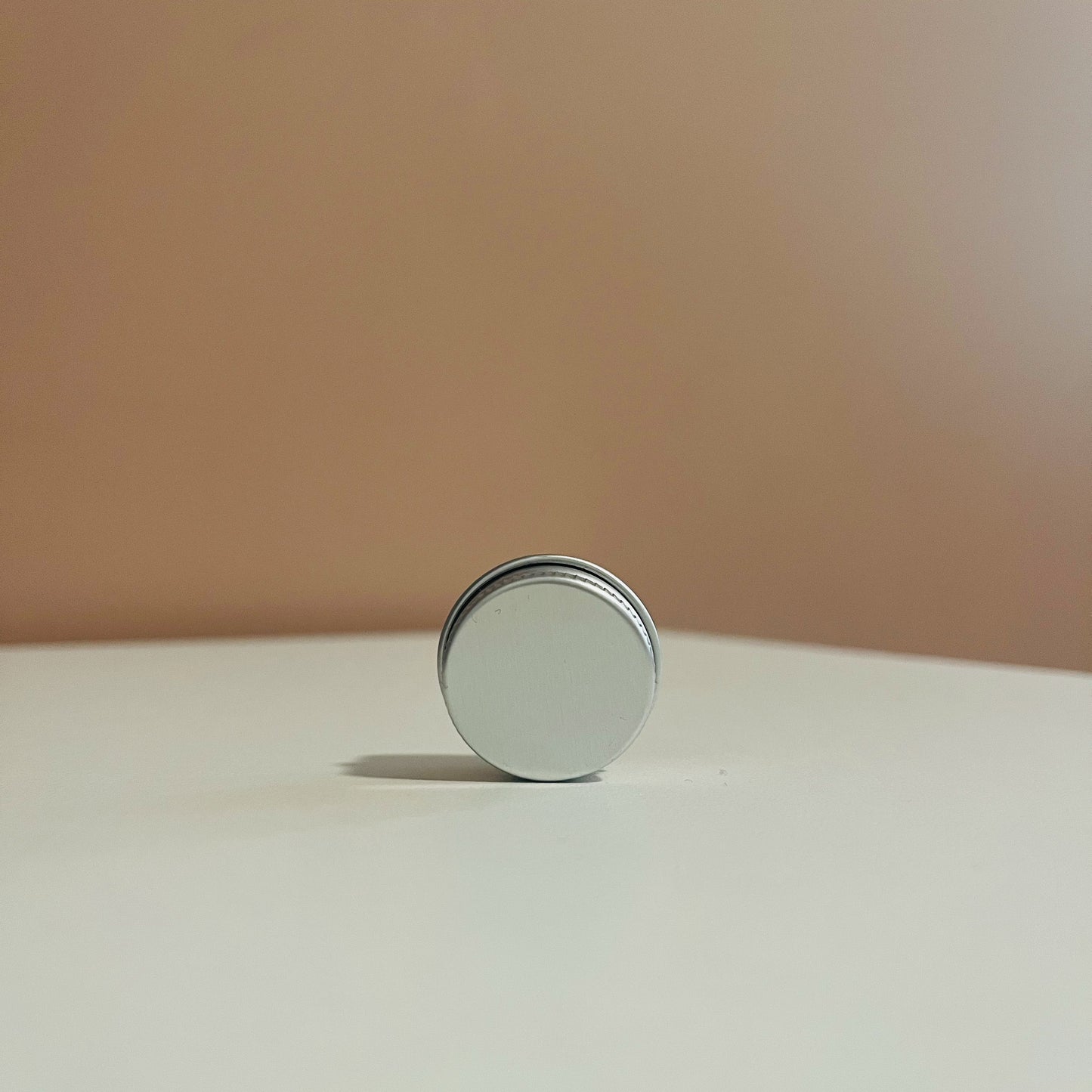 Silver cylindrical container filled with demulcere Salve lip balm and dry spot treatment on a beige surface with a beige background