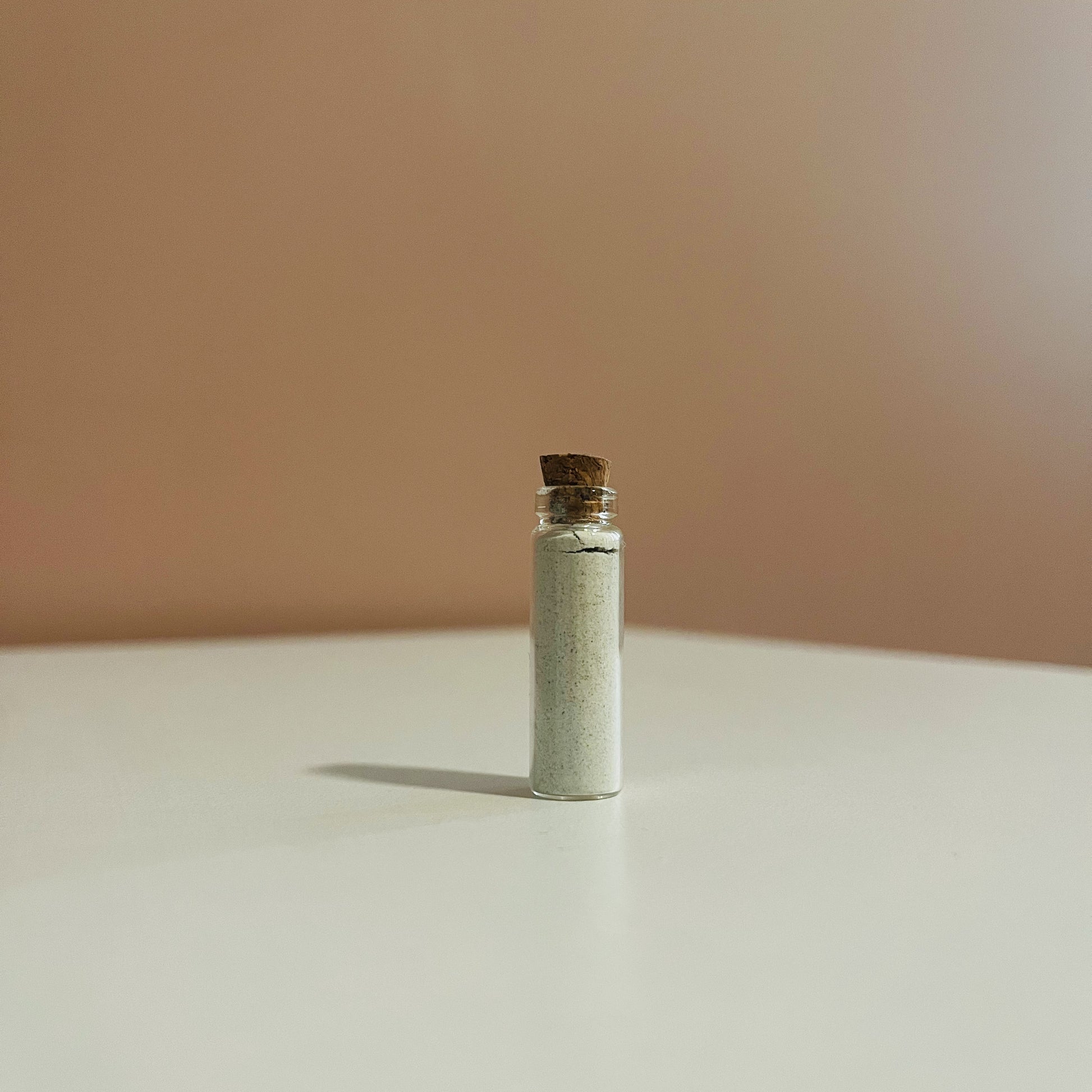 Small glass bottle filled with demulcere Suds powdered facial cleanser with cork lid on a beige surface and background