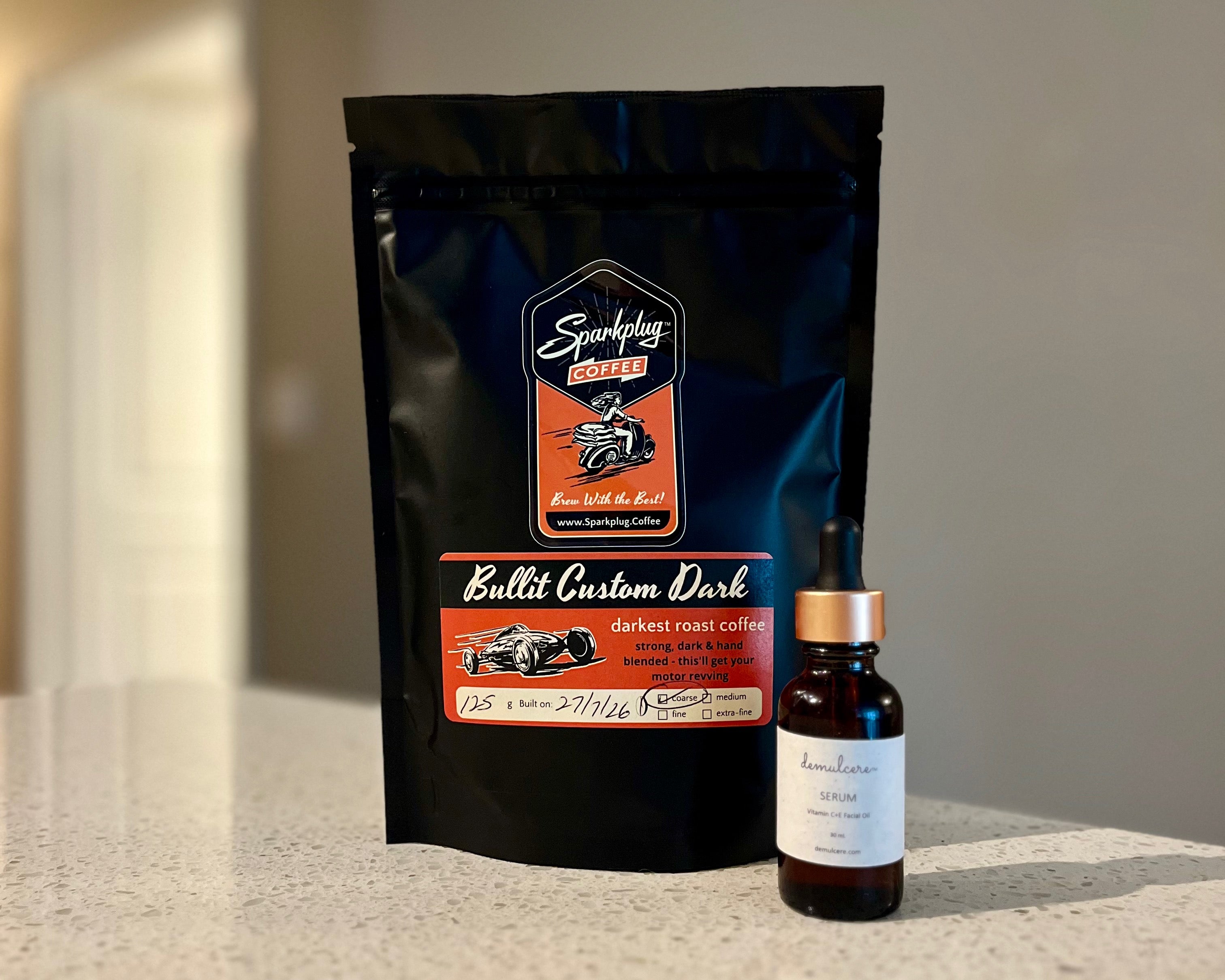 A black stand-up pouch of Sparkplug Bullit Custom Dark gourmet roast coffee stands on a light countertop next to an amber glass dropper bottle of Demulcere Serum with a white label and rose gold cap.